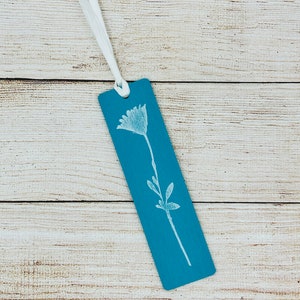May include: A teal blue bookmark with a white floral design and a white ribbon.