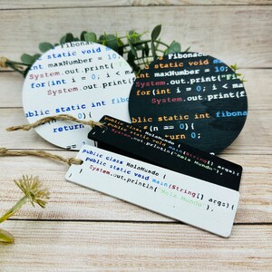 Op de afbeelding: Drie ronde houten ornamenten met geprinte code. De code is voor een "Fibonacci"-reeks, een "Hola Mundo"-programma en een "Fibonaco"-reeks. De ornamenten zijn wit, zwart en wit.