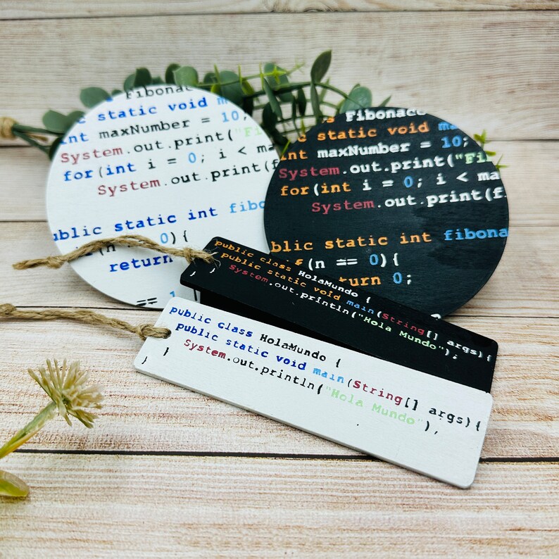 Op de afbeelding: Drie ronde houten ornamenten met geprinte code. De code is voor een "Fibonacci"-reeks, een "Hola Mundo"-programma en een "Fibonaco"-reeks. De ornamenten zijn wit, zwart en wit.