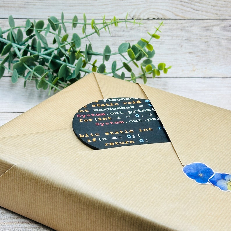Op de afbeelding: Een bruine papieren cadeauzak met een zwarte ronde sticker met een fragment van computercode. De code is in wit geschreven en bevat de woorden "Fibonace", "static", "void", "maxNumber", "System.out.print", "for", "int", "i", "0", "1", "System.out.pri", "blic", "static", "int", "f", "if", "n", "0", "return", "0".