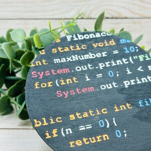 Op de afbeelding: Een ronde zwarte onderzetter met witte tekst die "Fibonaco c static void ma Int maxNumber = 10; System.out.print("Fil for(int i = 0; i < System.out.print blic static int fil if(n == 0) { return 0;" luidt.