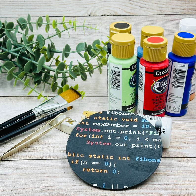 Op de afbeelding: Een ronde zwarte onderzetter met witte tekst die "Fibonacci static void main int maxNumber = 10; System.out.print("Fibonacci"); for(int i = 0; i < maxNumber; i++) { System.out.print(fibonacci(i)); } public static int fibonacci(int n) { if (n == 0) { return 0; } = 1 1 ||" luidt. De onderzetter is omringd door vijf flessen acrylverf in verschillende kleuren, waaronder rood, groen, blauw en zwart. Twee kwasten en een pincet zijn ook zichtbaar.