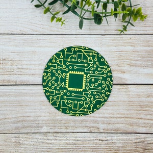 Puede incluir: Un posavasos redondo verde con un diseño de circuito impreso amarillo. El circuito impreso tiene un cuadrado central con un interior verde.