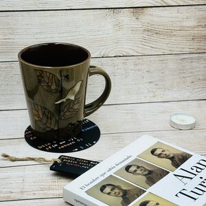 Op de afbeelding: Een bruine keramische mok met een bladontwerp en een kleine vogel op een tak. De mok staat op een zwarte onderzetter met witte tekst. De tekst op de onderzetter bevat codefragmenten en het woord "Fibonacci".