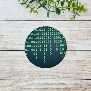 May include: A black round coaster with a green binary code pattern. The code includes the numbers 0 and 1.