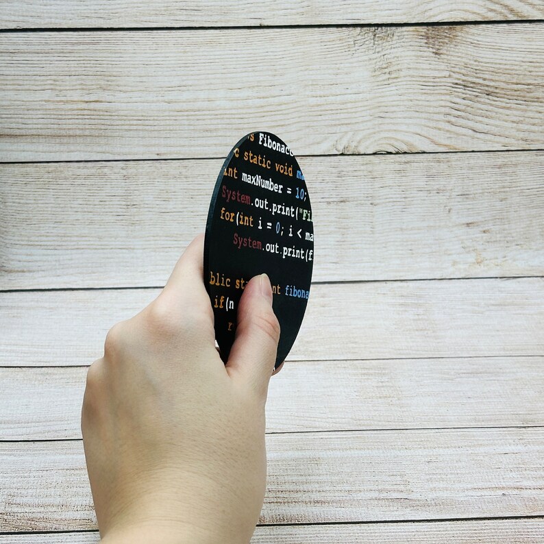 Op de afbeelding: Een ronde zwarte onderzetter met code erop gedrukt in verschillende kleuren. De code is voor een Fibonacci-sequentiefunctie. De onderzetter wordt vastgehouden door een hand.
