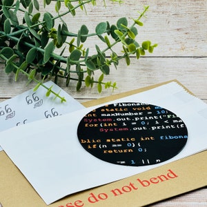 Op de afbeelding: Een ronde zwarte sticker met witte tekst die "Fibonaco static void main int maxNumber = 10; System.out.print("Fi for(int i = 0; i < ma System.out.print(f blic static int fibona if (n == 0) { return 0; = 1 || p" luidt. De sticker zit op een bruine papieren envelop met de tekst "Niet vouwen" erop gedrukt.