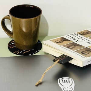 Op de afbeelding: Een bruine keramische mok met een zwarte onderzetter met code. Een boek met de titel "During Alan" ligt op een wit oppervlak met een bruin bladwijzer.