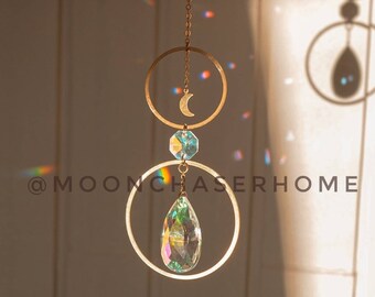 Puede incluir: Un atrapasol de cristal dorado con un dije de media luna y un cristal en forma de lágrima que cuelga de dos aros. El atrapasol proyecta arcoíris en una pared blanca.