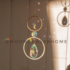 May include: A gold-toned crystal suncatcher with a crescent moon charm and a teardrop-shaped crystal hanging from two hoops. The suncatcher casts rainbows on a white wall.