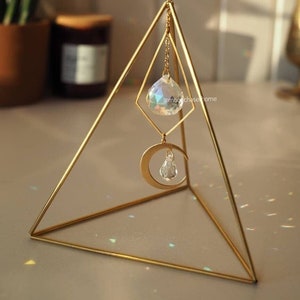 Puede incluir: Un soporte de pirámide geométrica de metal dorado con un prisma de cristal colgando de una cadena. El prisma es iridiscente y refleja la luz. El soporte está sobre una superficie blanca. @moonchasehome