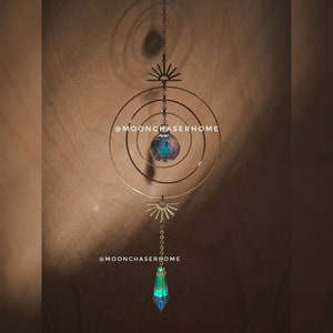 Può includere: Un design a forma di sole in metallo dorato con una sfera di cristallo appesa. La sfera di cristallo è iridescente e riflette la luce. Il sole è attaccato a una catena che pende dal soffitto. Il testo '@moonchaserhome' è visibile nell'immagine.