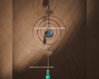 Puede incluir: Un diseño de sol en metal dorado con una bola de cristal colgando de él. La bola de cristal es iridiscente y refleja la luz. El sol está unido a una cadena que cuelga del techo. El texto '@moonchaserhome' es visible en la imagen.