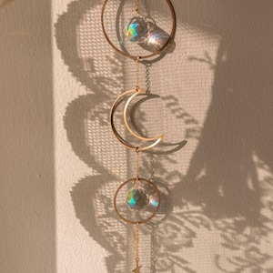 Pode incluir: Decoração suspensa com tema celestial com uma lua crescente dourada, duas bolas de cristal transparentes e dois círculos dourados. A decoração é suspensa por uma corrente dourada e inclui três pequenas estrelas douradas.