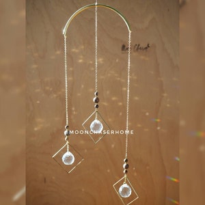 Puede incluir: Móvil colgante de metal dorado con tres esferas de cristal transparente suspendidas por delicadas cadenas. El móvil presenta una parte superior curvada y marcos cuadrados. El texto "MOONCHASERHOME" es visible.