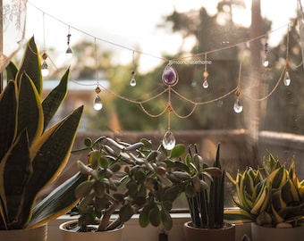 May include: A gold-colored chain with hanging crystals and gemstones, including a large amethyst, is suspended in front of a window. The crystals are teardrop-shaped and faceted. The text "moonchaserhome" is visible. Potted plants are in the foreground.