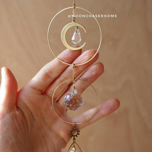 Puede incluir: Un atrapasol de cristal de sol y luna de metal dorado con una l&aacute;grima de cristal transparente que cuelga de la parte inferior. El atrapasol est&aacute; colgado de una cadena dorada.