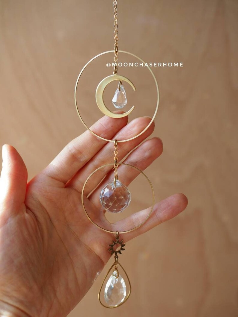Puede incluir: Un atrapasol de cristal de sol y luna de metal dorado con una l&aacute;grima de cristal transparente que cuelga de la parte inferior. El atrapasol est&aacute; colgado de una cadena dorada.