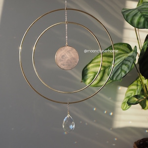 May include: A gold metal hoop mobile with a pink rose quartz crystal and a clear crystal teardrop hanging from it. The mobile is hanging in front of a green leafy plant.
