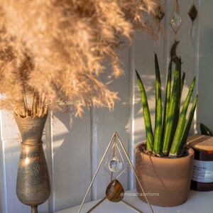 K&ouml;nnte beinhalten: Ein goldfarbener dreieckiger Metallst&auml;nder mit einem Kristallprisma und einem Tigerauge-Stein, der daran h&auml;ngt. Der St&auml;nder steht auf einer wei&szlig;en Oberfl&auml;che mit einer Vase mit getrocknetem Pampasgras und einer Topfpflanze im Hintergrund. @moochaserhome ist auf dem St&auml;nder geschrieben.