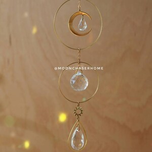 Puede incluir: M&oacute;vil colgante de metal dorado con sol, luna y prisma de cristal, con una l&aacute;grima de cristal transparente colgando de la parte inferior. El m&oacute;vil est&aacute; colgado de una cadena sobre un fondo marr&oacute;n claro.