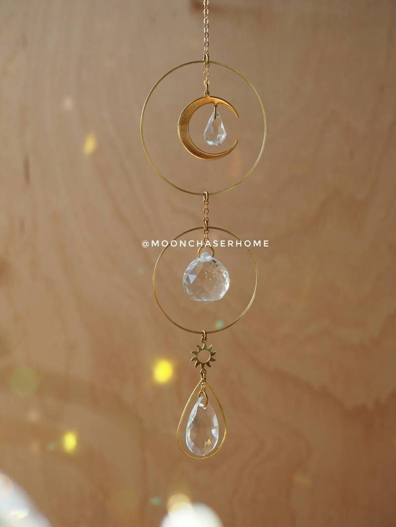 Puede incluir: M&oacute;vil colgante de metal dorado con sol, luna y prisma de cristal, con una l&aacute;grima de cristal transparente colgando de la parte inferior. El m&oacute;vil est&aacute; colgado de una cadena sobre un fondo marr&oacute;n claro.
