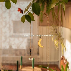 Puede incluir: Un atrapasoles de luna creciente dorada y cristal colgando de un anillo dorado, suspendido por delicadas cadenas. El atrapasoles presenta un cristal transparente y cuentas verdes y rojas. El texto "@MOONCHASERHOME" es visible.