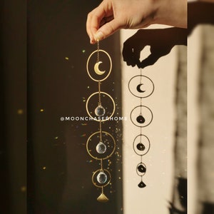 May include: A gold moon and crystal suncatcher with a crescent moon design and three crystal spheres hanging from a chain. The suncatcher is hanging from a hand and casting a shadow on the wall behind it. The text '@moonchaserhome' is visible in the image.