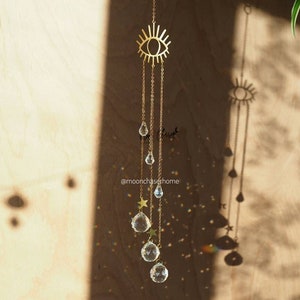May include: A gold eye-shaped suncatcher with crystal drops and star charms hanging from it. The suncatcher is hanging in front of a wooden wall with light shining through it. The text "@moonchaserhome" is visible at the bottom of the image.