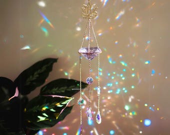 May include: A gold-toned crystal suncatcher with a lotus flower design at the top. The suncatcher has a large, clear crystal in the center and smaller crystals hanging from it. The suncatcher is casting rainbows on the wall behind it.
