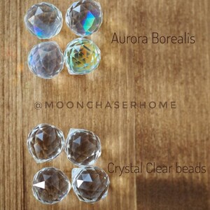 K&ouml;nnte beinhalten: Vier klare Glaskristallperlen, zwei mit einem schillernden Aurora-Borealis-Finish und zwei mit einem klaren Kristall-Finish. Die Perlen sind in zwei Reihen zu je zwei auf einer Holzoberfl&auml;che angeordnet. Der Text "Aurora Borealis" und "Kristallklare Perlen" steht unter den Perlen.