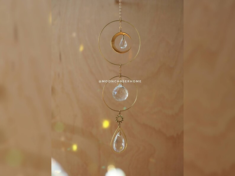 Puede incluir: Un m&oacute;vil colgante de metal dorado con tres prismas de cristal y un dije de luna creciente. El m&oacute;vil est&aacute; suspendido de una cadena y cuelga del techo.