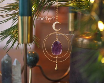 Op de afbeelding: Amethyst kristallen hangende ornament met een gouden halve maan en een dubbel cirkel ontwerp. De ornament hangt aan een gouden ketting en is omgeven door groen.