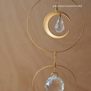 Puede incluir: Una luna creciente de metal dorado con una l&aacute;grima de cristal colgando de ella. La luna est&aacute; suspendida de una cadena y un anillo dorados. Debajo de la luna hay otro anillo dorado con un cristal m&aacute;s grande colgando de &eacute;l. El fondo es de color marr&oacute;n claro. @moonchaserhome