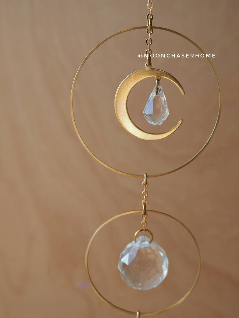 Puede incluir: Una luna creciente de metal dorado con una l&aacute;grima de cristal colgando de ella. La luna est&aacute; suspendida de una cadena y un anillo dorados. Debajo de la luna hay otro anillo dorado con un cristal m&aacute;s grande colgando de &eacute;l. El fondo es de color marr&oacute;n claro. @moonchaserhome