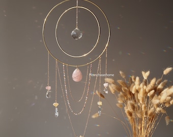 May include: A gold-colored metal dream catcher with a large, clear crystal ball and a teardrop-shaped rose quartz crystal. The dream catcher has multiple strands of crystals and beads hanging from it. The background is a neutral color.
