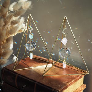Puede incluir: Dos soportes triangulares de metal dorado con atrapasoles de cristal colgando de ellos. Cada atrapasol tiene una bola de cristal, un colgante de cristal y un amuleto. Los amuletos son un sol y una luna creciente. Los soportes están sobre una pila de libros antiguos.