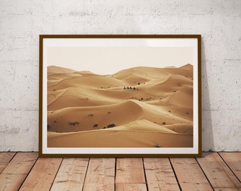 May include: A framed photograph of a desert landscape with rolling sand dunes. The dunes are a light brown color and there are a few small trees scattered throughout the scene. A group of camels is walking across the dunes in the distance.