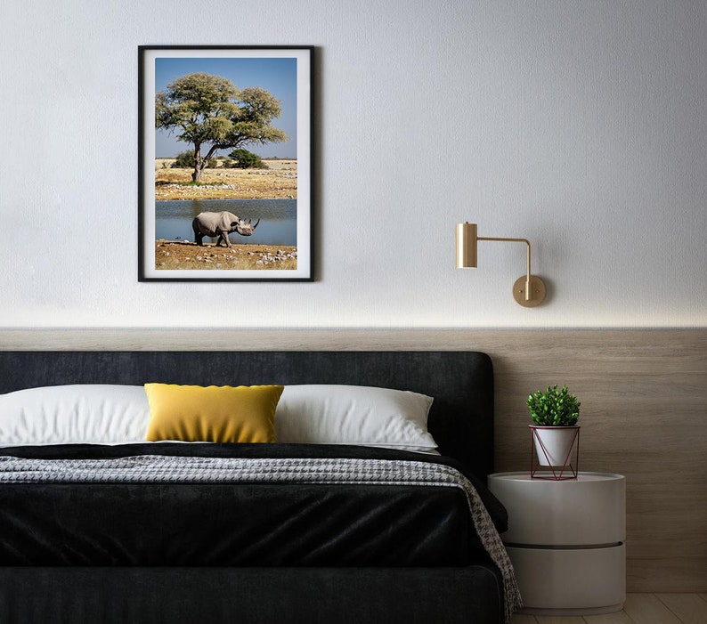 May include: A black and white framed photograph of a rhinoceros walking towards a body of water in a dry, grassy landscape. A tree with green leaves is in the background.