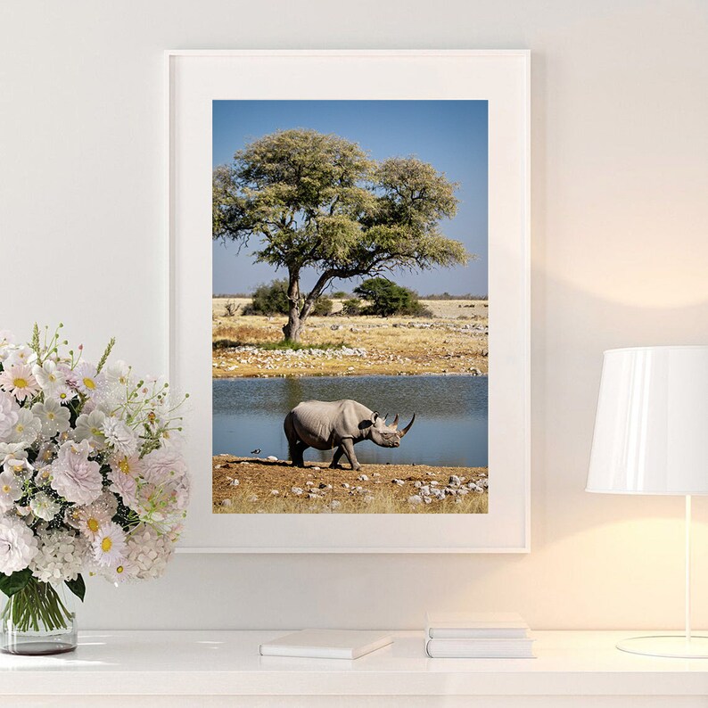 May include: A black rhinoceros walks along the edge of a watering hole in a savanna landscape. A large tree with green leaves stands in the background. The sky is blue and the water is clear.