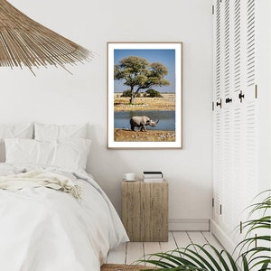 May include: A black rhinoceros walks towards a watering hole in a savanna landscape. A large tree with green leaves stands in the background. The image is framed in a light wood frame.