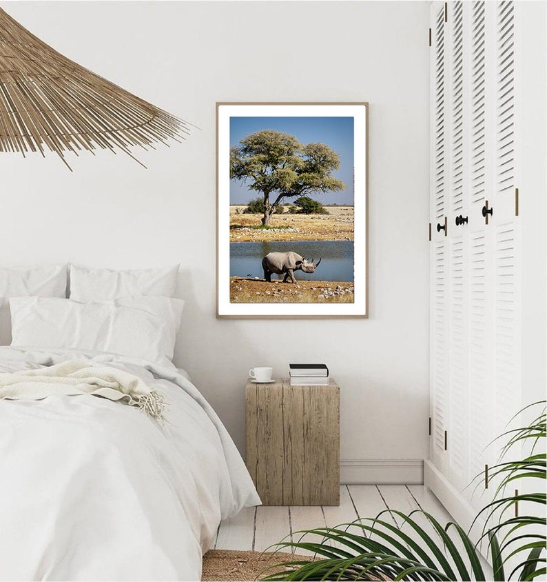 May include: A black rhinoceros walks towards a watering hole in a savanna landscape. A large tree with green leaves stands in the background. The image is framed in a light wood frame.