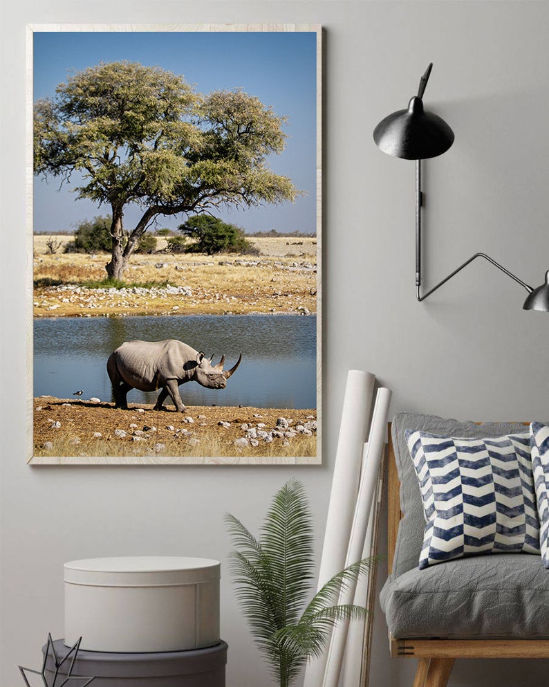 May include: A black rhinoceros walks towards a watering hole in a dry, grassy savanna. A large tree with green leaves stands in the background. The sky is blue and clear.