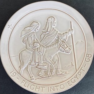 May include: Cream-coloured ceramic plate featuring a relief of a biblical scene. The image shows two figures, one riding a donkey, and another walking alongside. The text "19 - FLIGHT INTO EGYPT - G8" is inscribed around the edge, with a star in the upper left.