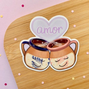 May include: Two brown ceramic mugs with blue and pink floral designs, with steam rising from them, under a purple heart with the word "amor" written in white.