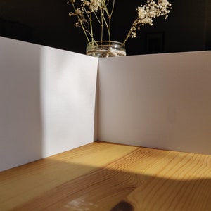 May include: A blank white card folded in half, sitting on a wooden table. The card is illuminated by sunlight, casting a shadow on the table.