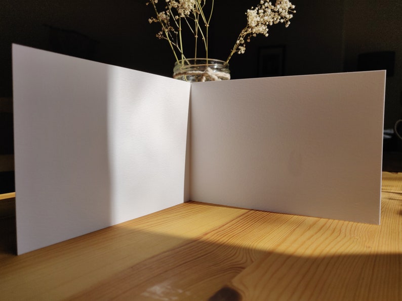 May include: A blank white card folded in half, sitting on a wooden table. The card is illuminated by sunlight, casting a shadow on the table.