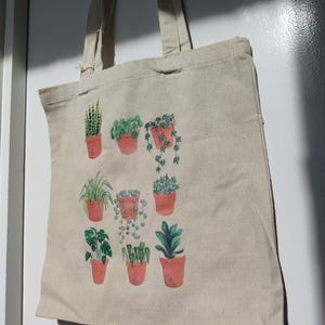 May include: A natural-coloured tote bag with a watercolour design of potted plants. The plants, in terracotta pots, include snake plants and trailing vines. The bag has long handles. The design is a collection of different green plants.