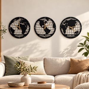 Puede incluir: Tres relojes de pared circulares de metal negro con un diseño de mapa mundial y líneas de cuadrícula. Cada reloj tiene manecillas doradas y los nombres de las ciudades LONDON, NEW YORK y TOKYO en letras mayúsculas blancas en la base.