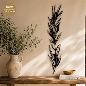 May include: A black metal wall art piece shaped like an olive branch with leaves and fruit, measuring 70 cm in height. The design features a vertical stem with multiple elongated leaves and several round olives arranged along the branch.